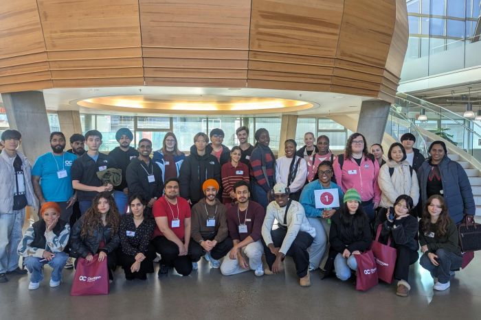 A group of people posing together indoors in front of a large wooden structure with curved panels and a staircase in the background.
