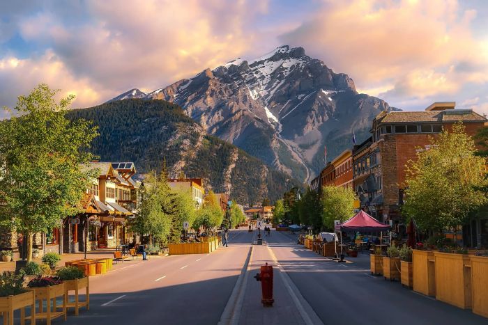 A quiet town street lined with wooden planters and buildings leads toward a massive, snow-capped mountain under a soft, golden sunset sky in Banff.