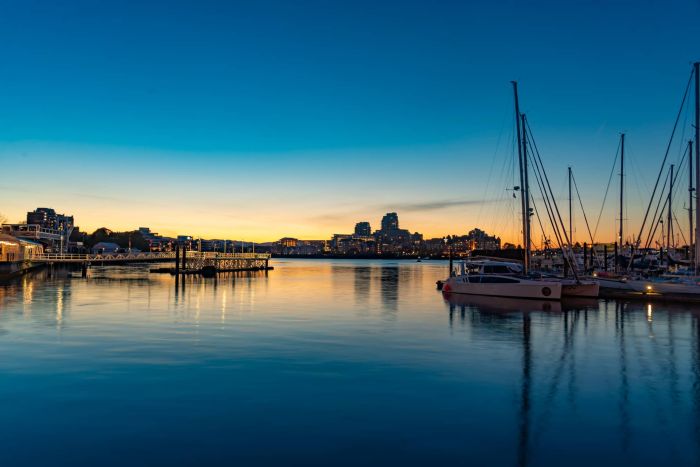 Victoria harbor at sunset with sailboats moored on the right, a pier on the left, and a distant city skyline reflecting on the calm, dark blue water.