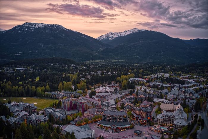 An aerial view of a Banff nestled in a valley with snow-capped peaks under a soft, pink and purple sunset sky surrounded by pine forests.