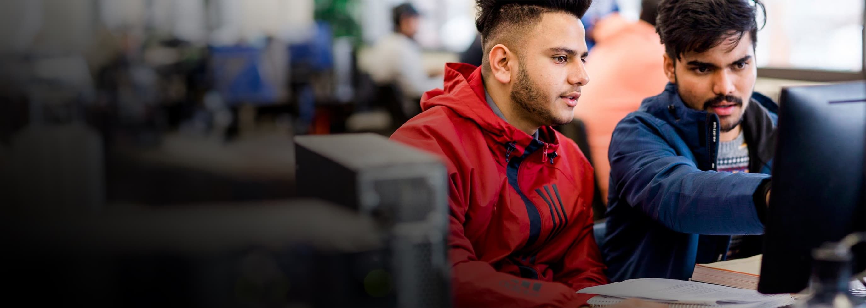 two students working on a desktop computer with one of the students pointing to the monitor and explaining something