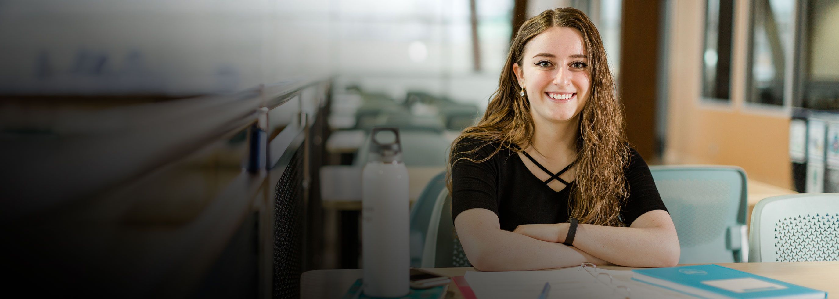 A smiling student with long wavy hair sits at a study table with open binders and notebooks in a bright, modern campus space.