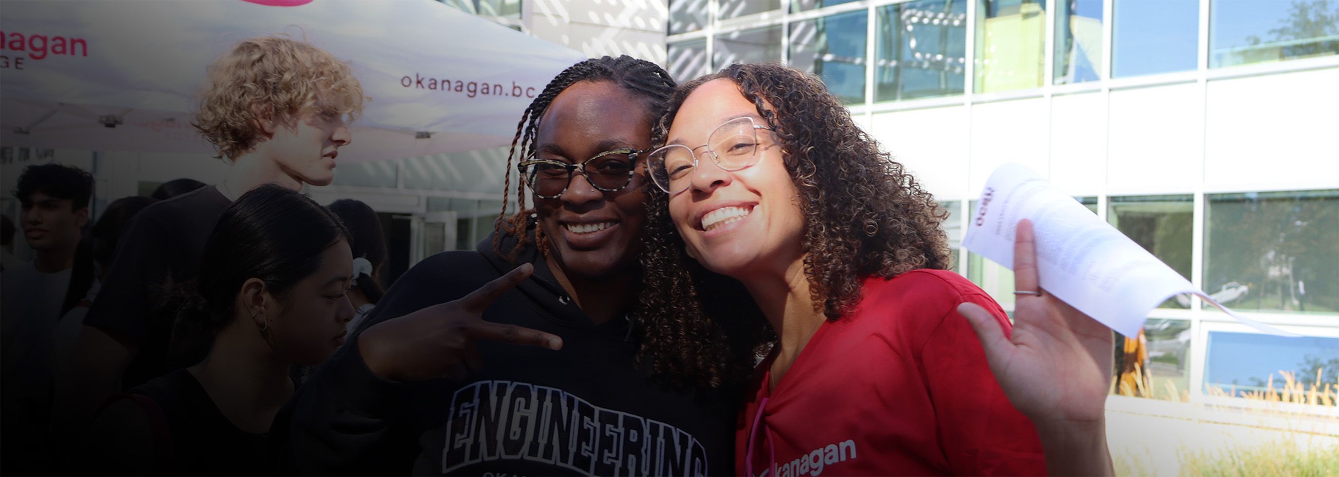 Two female students smiling and waving to the camera at an outdoor orientation event at the Kelowna campus