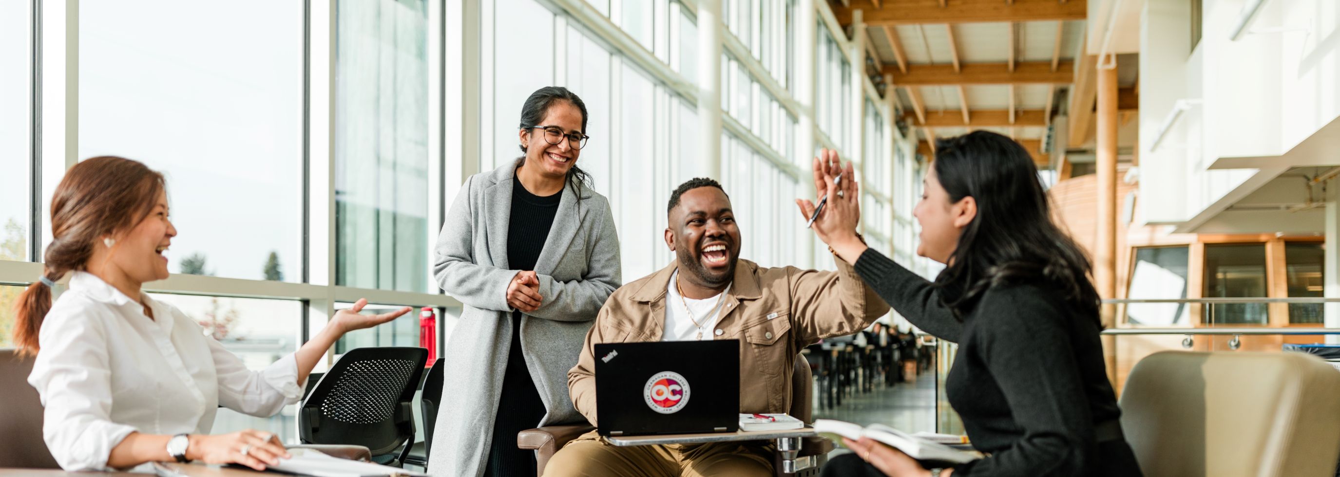 Four students gathered in a bright, modern campus lounge, sitting in a circle of armchairs with notebooks and a laptop. One student, seated with a laptop displaying the Okanagan College logo, is smiling and giving a high five to another student across from him. Two other students are laughing and engaged in the conversation, creating a warm, collaborative atmosphere.