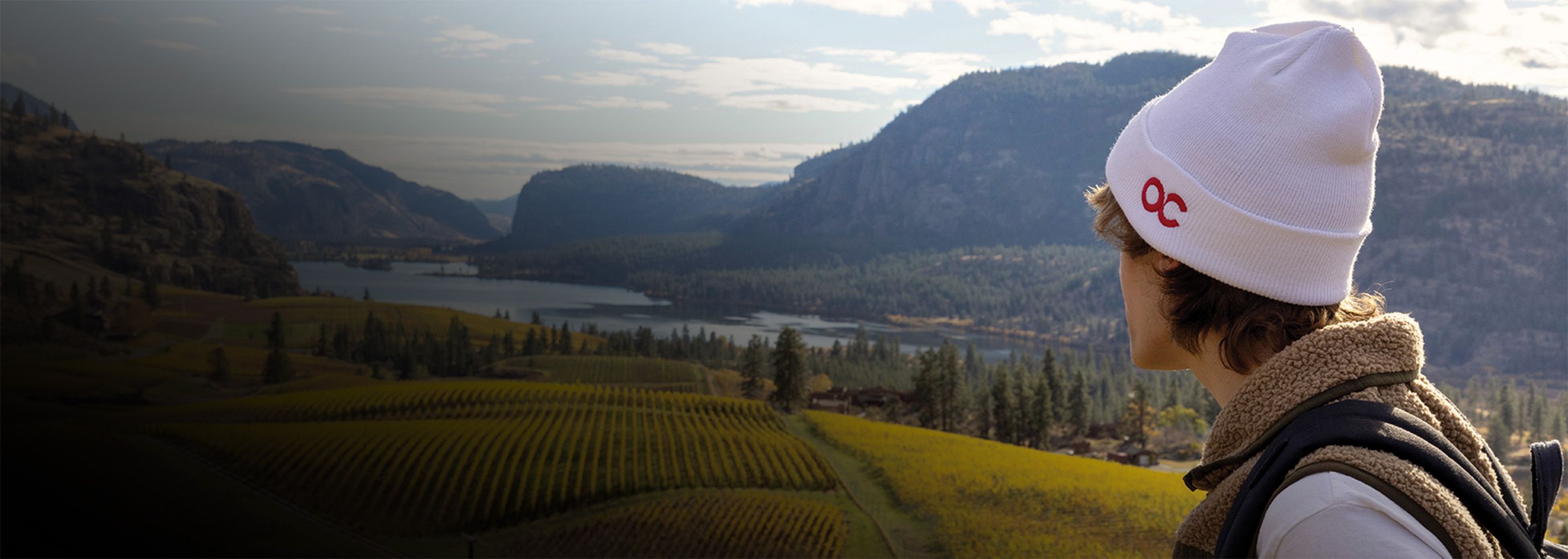 a person standing on a hill wearing a hat with Okanagan College icon and gazing at a serene view of vinyards and lake Okanagan