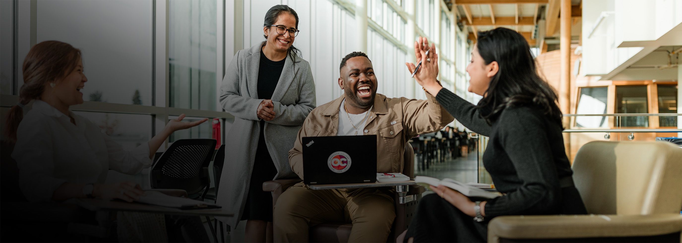 Four people are sitting in a modern, well-lit indoor space with large windows; one person is using a laptop, and two others are giving each other a high-five.