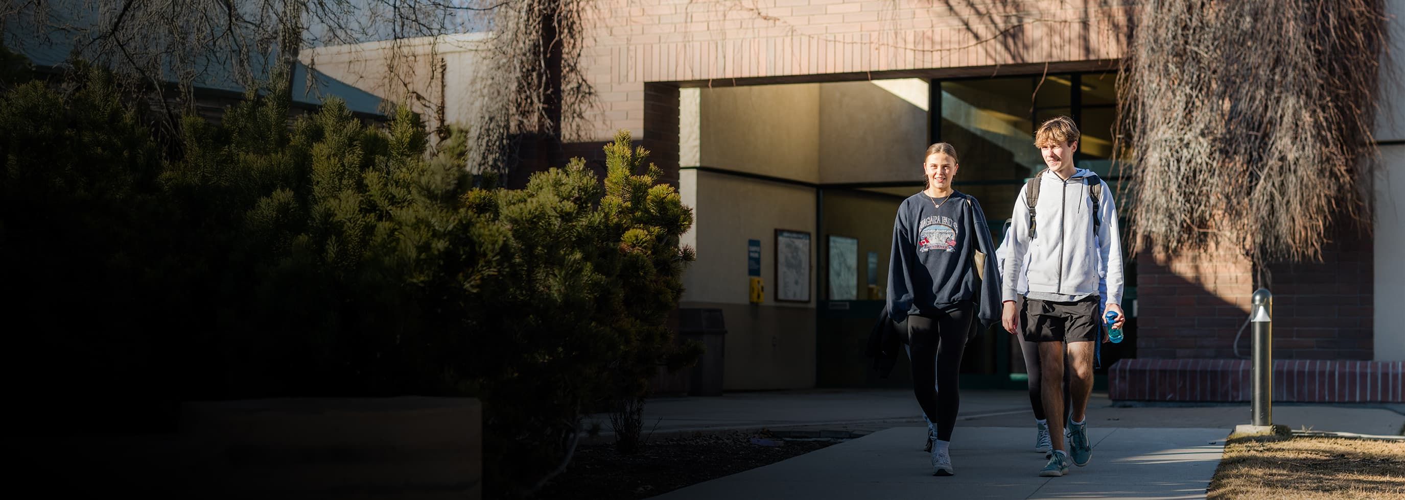  OC students walk on a sidewalk outside a building, carrying backpacks and wearing casual clothes on a sunny day.