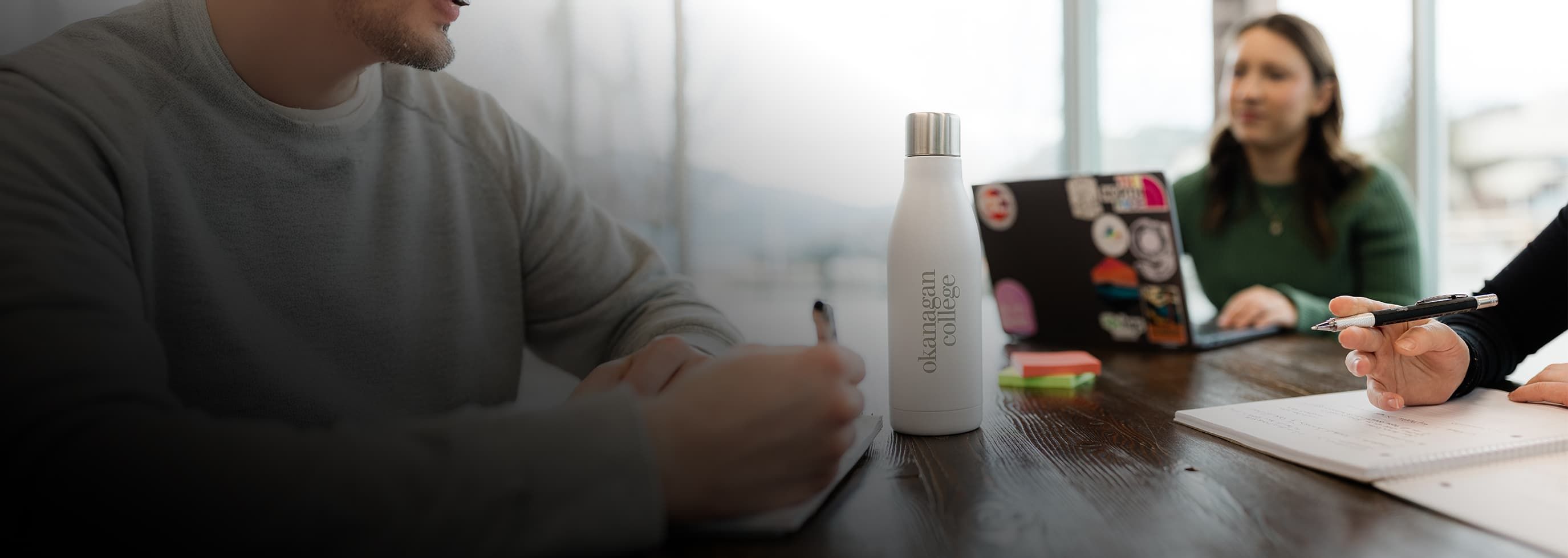 OC students siting at a table, writing in a notebook, engaged in conversation.