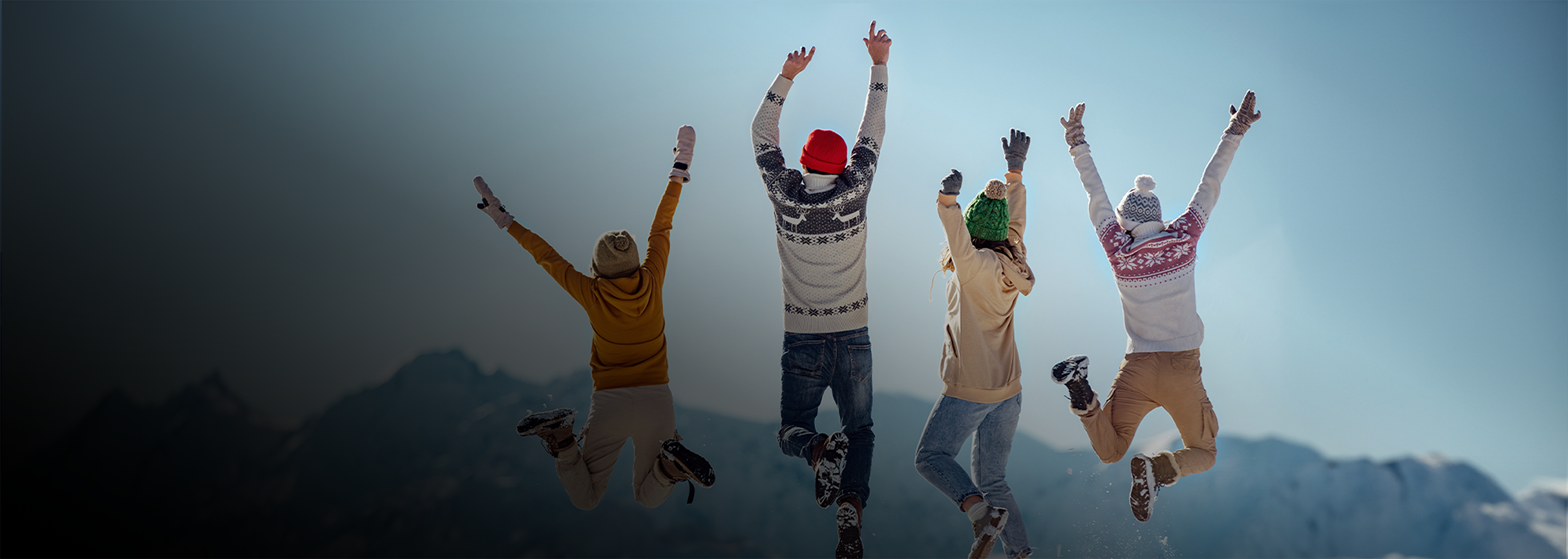Four people wearing winter clothing and colorful hats jump in the air against a backdrop of snow-covered mountains