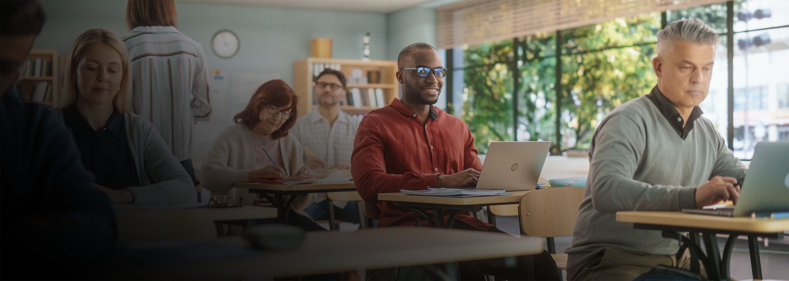 People sit at desks in a classroom using laptops and notebooks.