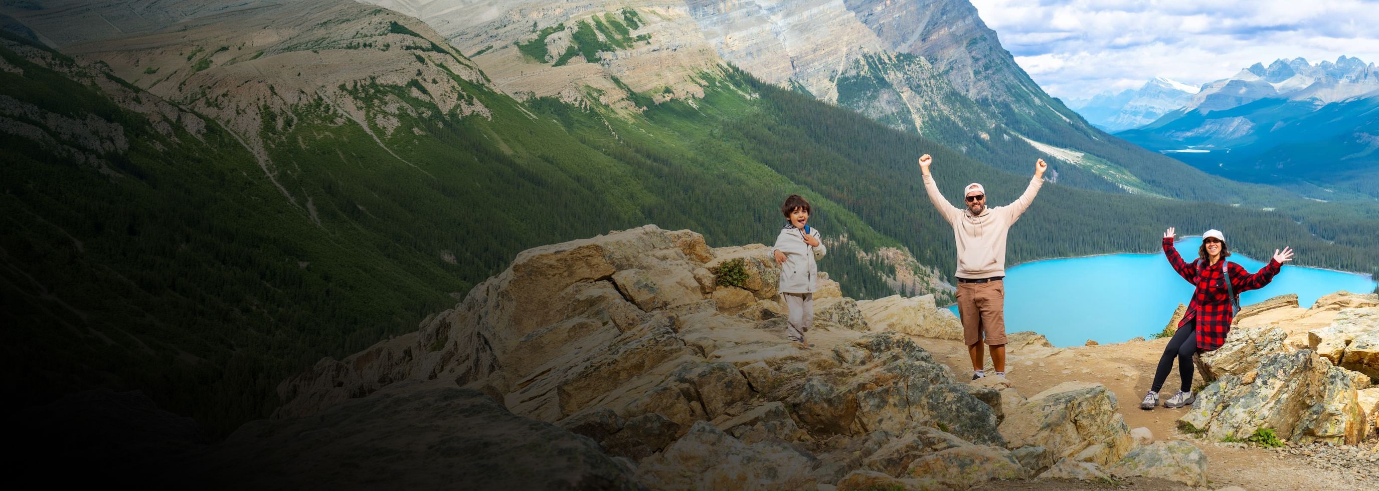 Three people posing on a rocky mountain overlook with a bright turquoise lake and vast forested mountains in the background under a blue sky.