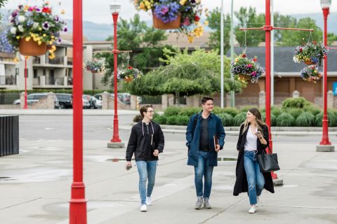 Two male students and one female student walking onto campus. Red posts are decorated with floral hanging baskets.