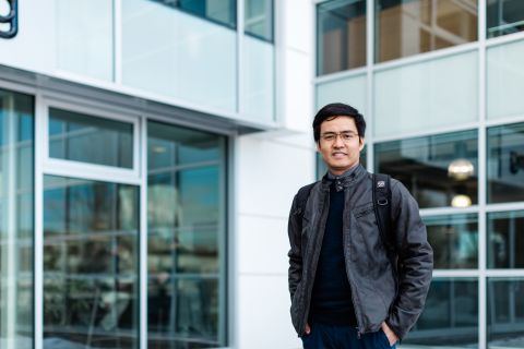 An international male student wearing a leather jacket and glasses stands outside in front of a building with a lot of windows.