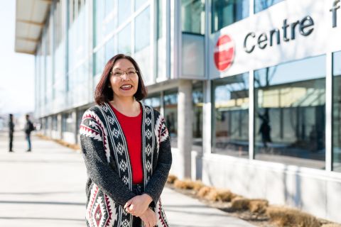 An Indigenous female student stands outside in front of the Centre for Learning building wearing red.