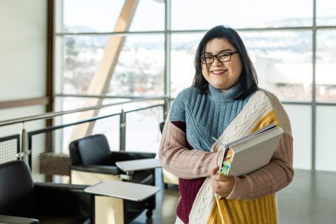 A female international student stands in front of an open study area holding books and wearing glasses.