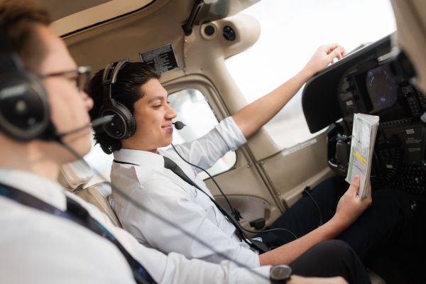 Two individuals wearing headsets and white shirts are seated in an aircraft cockpit, with one holding a folded map and the other adjusting a control near the instrument panel.