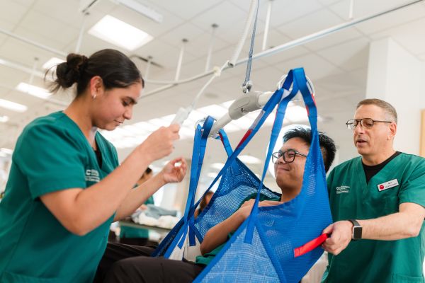 Two people in green uniforms are assisting an individual seated in a blue patient lift sling attached to an overhead hoist in a clinical setting.