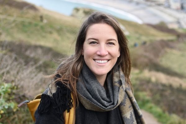 A female stands on a hill outside in a jacket and scarf wearing a backpack.