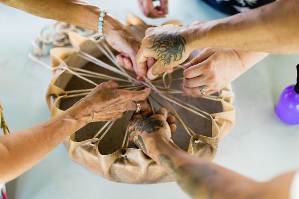 Five hands holding a hand drum upside down tying the hide on the drum. 