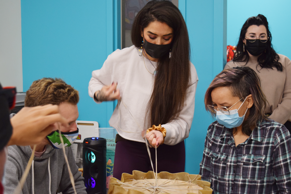 A person with long dark hair is teaching two students how to lace their hand drum