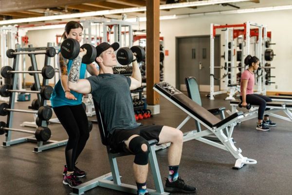 Student spotting another OC Kinesiology student lifting dumbell weights in a gym