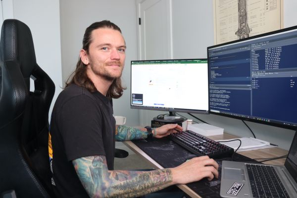 ELEN graduate works remotely from home, sitting at his computer desk with multiple screens.