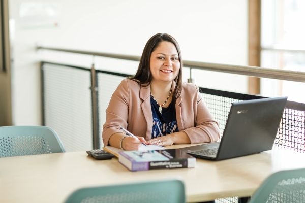 a smiling person working behind the desk with notes, calculator and and open laptop