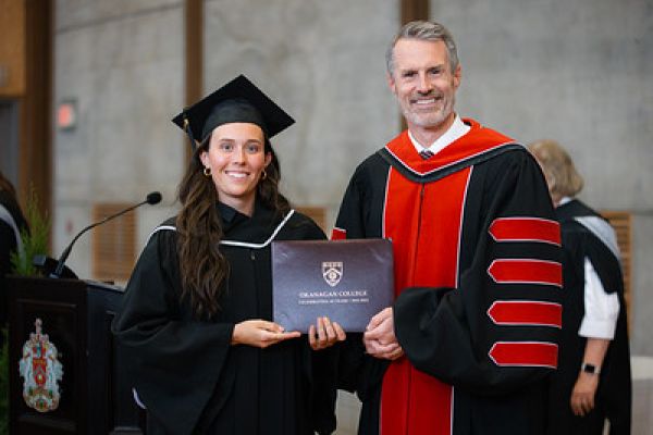 Female student in graduation robe and hat holding diploma next to College president at Convocation