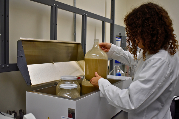 Student researcher in lab coat holding up glass jar of liquid in lab