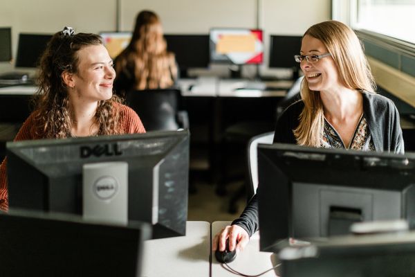 Two female students at computer stations smiling at each other