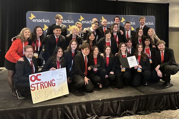 Enactus group photo. Students smiling together while on stage holding a sign that reads "O C strong, all day long"