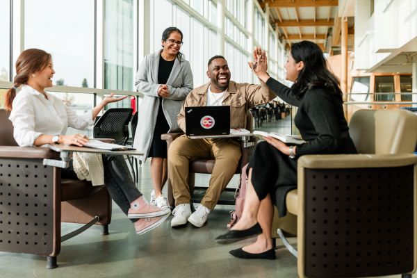 Four students gathered in a bright, modern campus lounge, sitting in a circle of armchairs with notebooks and a laptop. One student, seated with a laptop displaying the Okanagan College logo, is smiling and giving a high five to another student across from him. Two other students are laughing and engaged in the conversation, creating a warm, collaborative atmosphere.