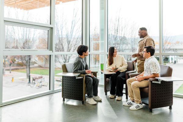 Four college students sit and stand in a bright, window-lined common area, engaged in a friendly conversation. One student is standing while the others are seated in study chairs with attached desks. Trees and campus buildings are visible outside the large windows. The atmosphere is casual and welcoming.
