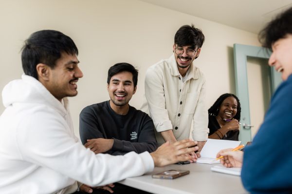 A group of five college students sit and stand around a table, smiling and laughing together during a casual study or discussion session. Notebooks and a phone are on the table, and the mood is friendly and collaborative.