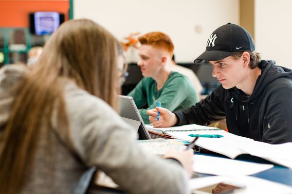 Three students are sitting at a table, working on their laptops and notebooks in a study environment.