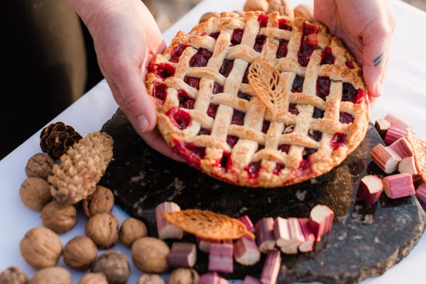 a lattice-topped pie above a black stone platter decorated with walnuts, rhubarb pieces, and pinecones.