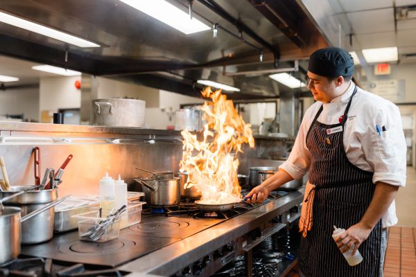 A chef flambés a dish in a commercial kitchen, sending flames rising dramatically from the pan.