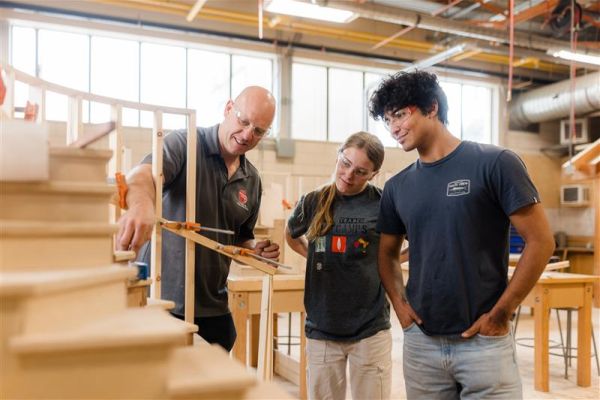 Three people in a woodworking workshop, with one demonstrating a measuring tool near a wooden staircase while the others observe.