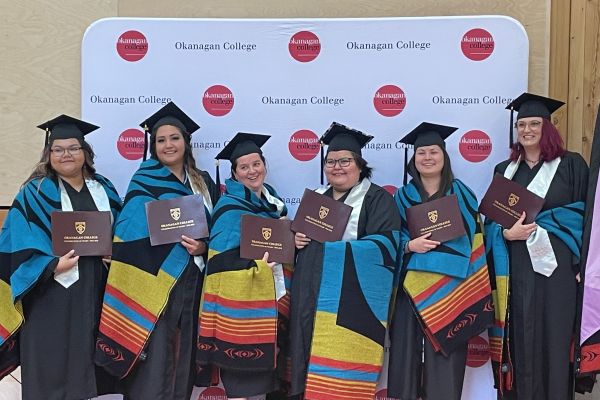 Group of graduates in caps and gowns holding diplomas and wearing colorful ceremonial blankets, standing in front of an Okanagan College backdrop during a graduation event