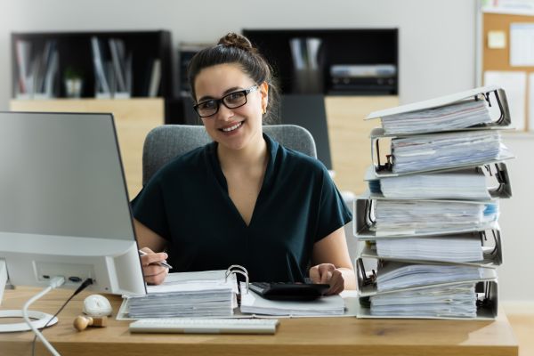 Person seated at a desk in an office environment working on paperwork with a computer monitor and a large stack of binders