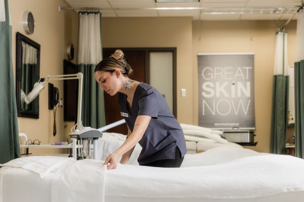 Person arranging a treatment bed in a spa-like classroom setting with skincare equipment and a poster reading ‘Great Skin Now,