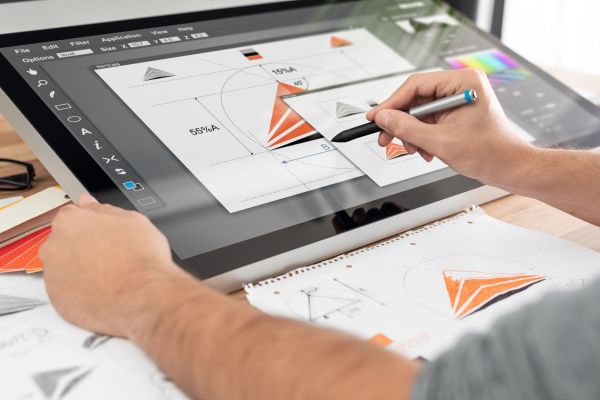 Person using a stylus on a large digital drawing tablet to create geometric designs and color layouts, with sketches and notes on paper nearby