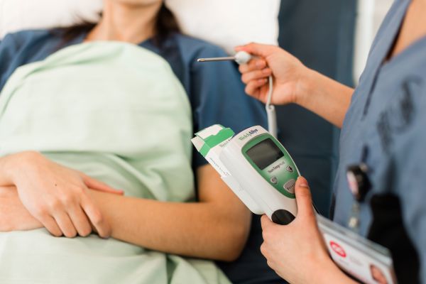 Healthcare professional holding a digital thermometer and a vital signs monitor while attending to a patient lying under a green blanket