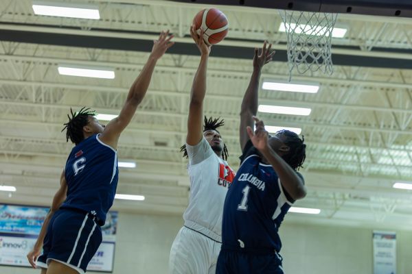 Okanagan College men’s basketball game showing a player in a white uniform attempting a layup while two defenders in blue Columbia uniforms contest the shot near the hoop.