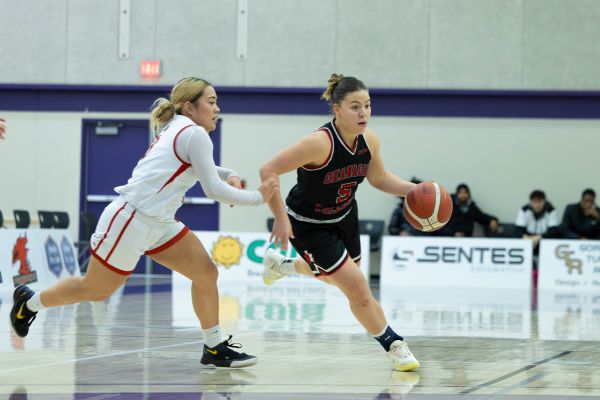 Okanagan College women’s basketball game showing a player in a black uniform dribbling the ball while being closely defended by an opponent in a white uniform on an indoor court.