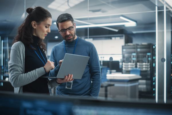 Two individuals standing in a modern server room reviewing information on a tablet, with racks of network equipment visible in the background