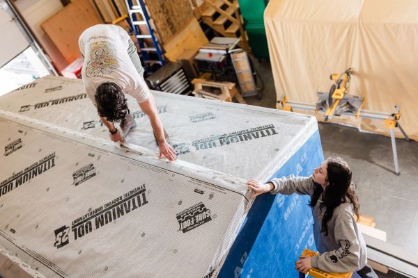 Two people affix synthetic roofing underlayment to a structure in a workshop. One is on top working, while the other assists from the side.
