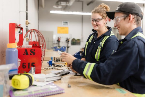 Two individuals wearing dark work uniforms with reflective safety stripes are seated at a workbench, reviewing a small mechanical part and paperwork, with tools, notebooks, and equipment visible on the table in an industrial workshop setting.