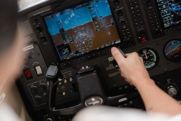 A close-up view of an aircraft cockpit showing a hand on the control yoke with digital flight instruments and navigation displays on the panel.
