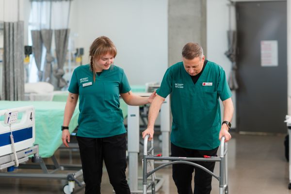 Two people wearing green uniforms are in a clinical setting, with one person assisting the other who is using a walker near hospital beds.